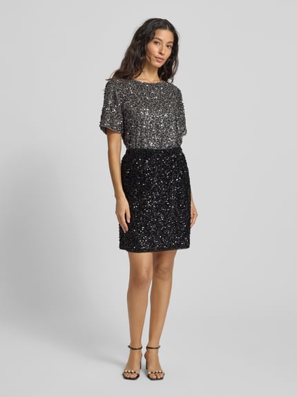 Frauenmodel trägt Minirock mit Pailletten und elastischem Bund von comma, schwarze Glitzertextur, gerader Schnitt, Ganzkörperansicht von vorne.
