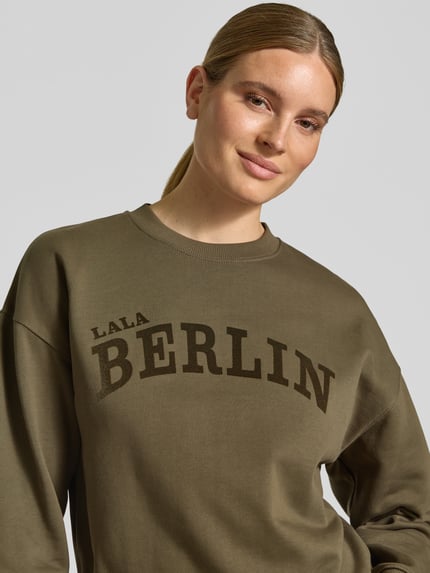 Frauenmodel trägt Sweatshirt mit Label-Print von Lala Berlin in Olivgrün mit Rundhalsausschnitt und gerippten Abschlüssen, Nahaufnahme Oberkörper.