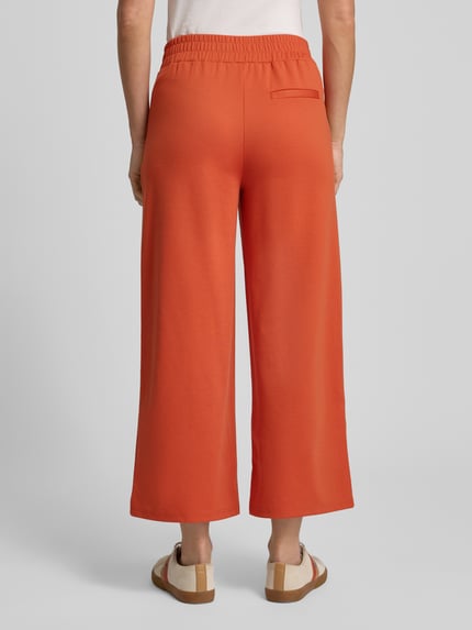 Kobiece modelka nosi spodnie materiałowe Wide Fit Stoffhose mit Viskose-Anteil Model 'Kate' od ICHI, elastyczna mieszanka materiałów z dodatkiem wiskozy, szerokie nogawki, kieszeń z tyłu, zakładki w pasie, elastyczny pas, pomarańczowy kolor, faktura gładka, widok całościowy z tyłu.
