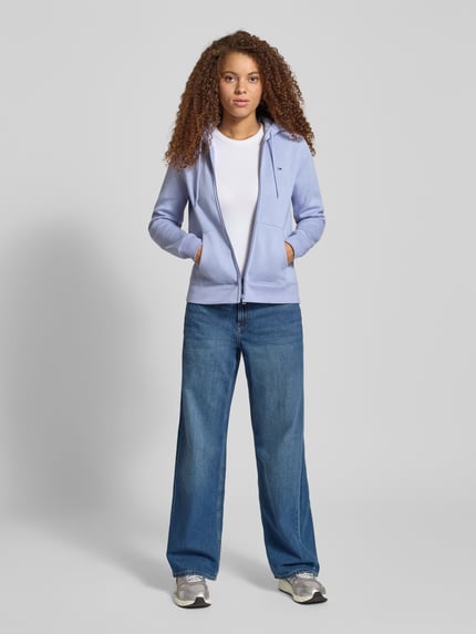 Kobiece modelka nosi Regular Fit Sweatjacke aus Baumwoll-Mix od Tommy Jeans w kolorze jasnofioletowym, z kapturem i zamkiem, bawełniany materiał, przód w pełnej długości, unikalny detal logo, widok całej sylwetki z przodu.