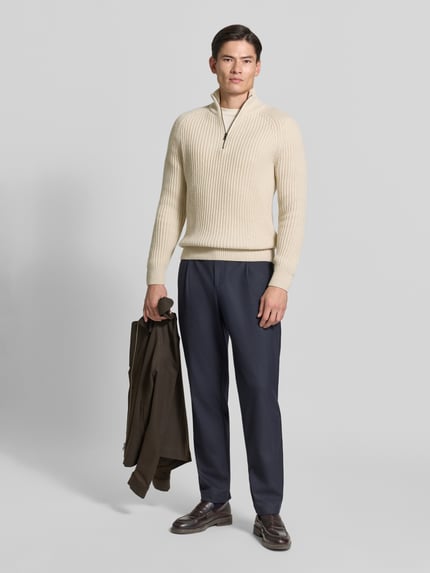 Męski model nosi sweter z dzianiny 'Strickpullover aus Schurwoll-Mix mit Troyer-Kragen' od ROY ROBSON, mieszanka żywej wełny z domieszką wełny z alpaki, krój slim fit, Kołnierz zapinany, detal z logo, długość tyłu w rozmiarze M: 78 cm, długość rękawów w rozmiarze M: 80 cm, kolor kremowy, prążkowana faktura, spodnie granatowe, widok całej sylwetki z przodu.