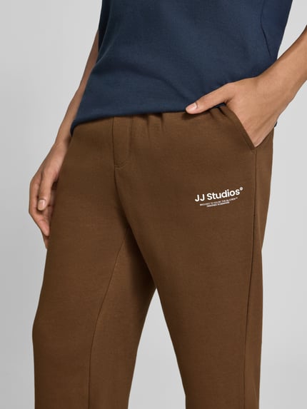 Männermodel trägt Straight Fit Sweatpants aus mieszanka bawełny Model 'Kane' od JACK & JONES, detal z logo, kieszeń z tyłu, elastyczny pas, elastyczne zakończenia nogawek, kolor brązowy, Długość wewnętrzna nogawki w rozmiarze M: 72 cm, zbliżenie na biodra