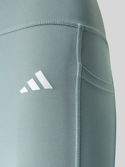 Frauenmodel trägt Leggings mit Logo und elastischem Bund von adidas Sportswear in grün, Stretch-Material, Nahaufnahme Oberschenkel.
