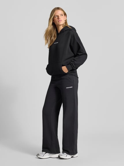 Frauenmodel trägt Wide Fit Sweatpants aus Baumwoll-Mix Modell 'ANNI' von PEGADOR in Schwarz mit Label-Detail, Ganzkörperansicht von vorne.