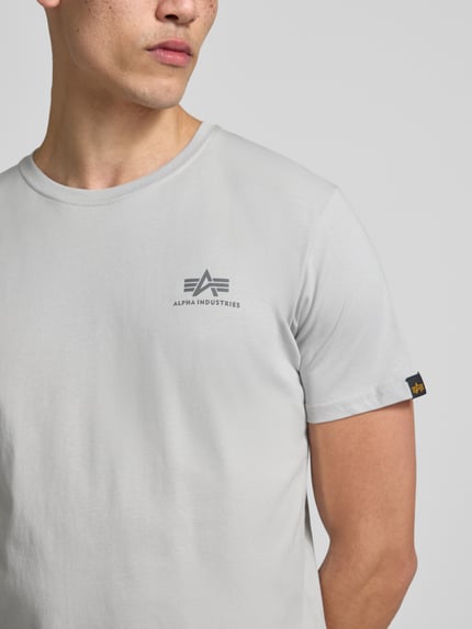 Männermodel trägt T-shirt mit Label-Details Modell 'BASIC' van Alpha Industries, puur katoen, recht model, ronde hals, ruglengte maat M 67 cm, nahaufnahme bovenlichaam.