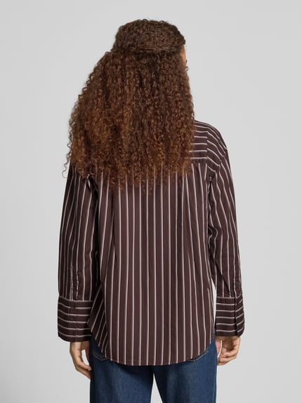 Männermodel trägt Oversized Hemdbluse mit Brusttasche von Gina Tricot z bawełny, krój oversized fit, kołnierzyk typu kent, listwa guzikowa, brustasza, sportowe mankiety, zaokrąglony dół, długość tyłu w rozmiarze S 71 cm, długość rękawów w rozmiarze S 54 cm, ciemnobrązowe tło z białymi paskami, faktura gładka, widoczne pionowe linie, Ganzkörperansicht Rückansicht