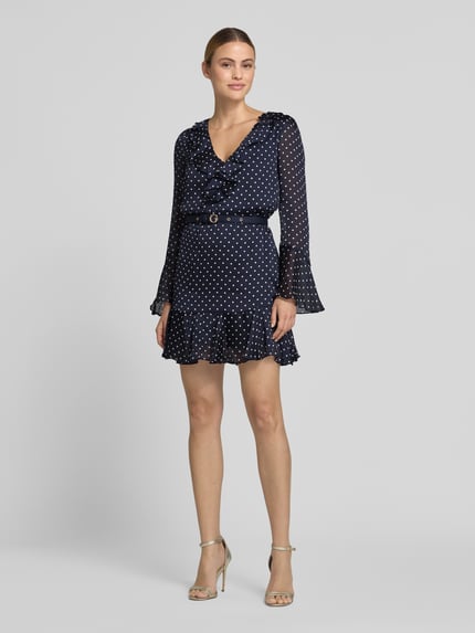 Frauenmodel trägt Minikleid mit Rüschenbesatz von GUESS, Tailliert, Abnehmbarer Taillen, V-Ausschnitt, Langärmelig, Punkte-Muster, Ganzkörperansicht von vorne. Rückenlänge bei Größe S: 83 cm, Brustweite bei Größe S: 42 cm, Ärmellänge bei Größe S: 62 cm.