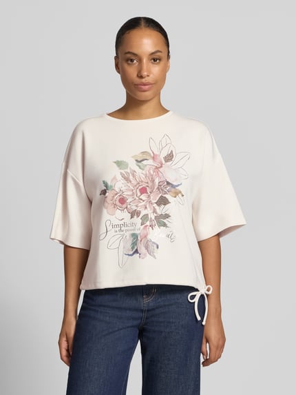 Frauenmodel trägt Relaxed Fit T-Shirt mit Statement-Print von Christian Berg Woman, florales Muster, helles Rosa, weich fallender Stoff, Ganzkörperansicht von vorne. Damen T-Shirt, elastischer Material-Mix mit Modal-Anteil, 1/2-Arm.