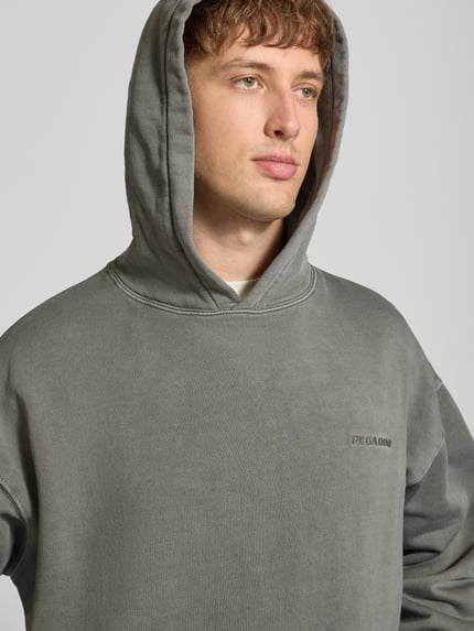 Männermodel trägt Oversized Hoodie aus Baumwoll-Mix von PEGADOR in Grau mit Logo-Stickerei und überschnittenen Schultern, Nahaufnahme Oberkörper.