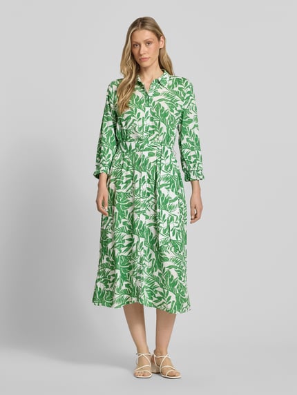 Modelka kobieca ma na sobie „Midikleid aus reiner Viskose mit Umlegekragen” od More & More, zielono-biały, florystyczny print, krój taliowany, pasek, półrękaw, guziki na całej długości, elastyczne mankiety, długość tyłu w rozmiarze 36: 125 cm, szerokość w rozmiarze 36: 40 cm, długość rękawa w rozmiarze 36: 49 cm; widok całej sylwetki z przodu.