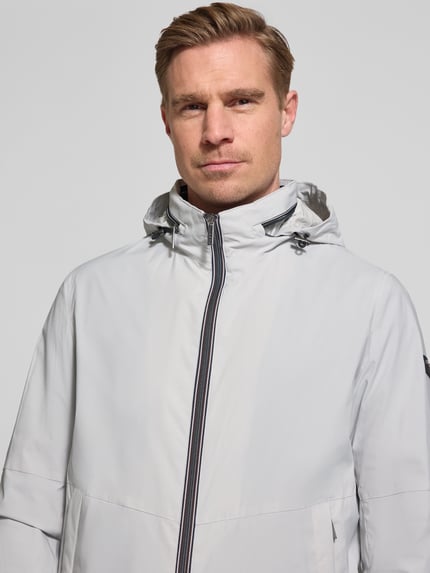 Männermodel trägt Regular Fit Jacke mit Stehkragen von bugatti in hellem Grau, mit Logo-Detail, verstaubarer Kapuze, Zweiwege-Reißverschluss, Eingrifftaschen und elastischen Ärmelabschlüssen, Nahaufnahme Oberkörper. Rückenlänge 68 cm, Schulterbreite 42 cm, Brustweite 50 cm, Ärmellänge 65 cm bei Größe 50.