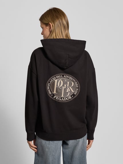 Vrouwenmodel draagt zwarte Oversized Hoodie met kangoeroezak model 'AVERSA' van Pegador, katoenmix, logo, rugaanzicht.
