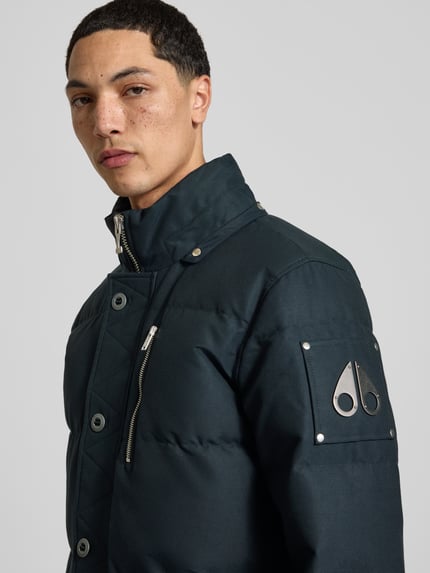 Männermodel trägt Daunenjacke mit abnehmbarer Fellkapuze von Moose Knuckles, unifarbene Webware, Struktur, Logo-Patch, Nahaufnahme Oberkörper.