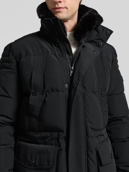 Mannenmodel draagt lange zwarte unikleuren Parka met capuchon van NAUTGÅRD 'J-LHOTSE 226', label-detail, sierzak, deelnaden, rits, drukknoopsluiting, steekzakken, ritszakken, binnenzak, elastische lange mouwen, tunnelkoord, detailopname bovenlichaam.