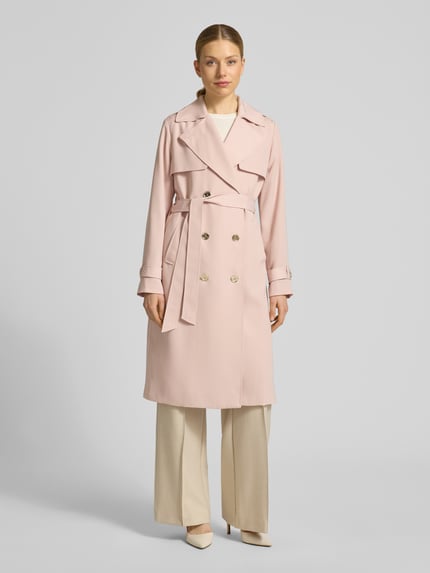 Vrouwenmodel draagt Zweireihiger Trenchcoat mit Bindegürtel van MICHAEL Michael Kors in lichtroze, double-breasted, reverskraag, regular fit, met riem, beige broek en pumps, lichte achtergrond. Volledig lichaamsaanzicht van voren.