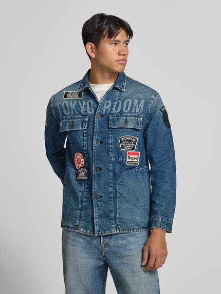 Männermodel trägt Regular Fit jeansjack van katoenmix met all-over labelpatches, borstzakken, steekzakken en platte kraag, knoopsluiting, Regular Fit Jeansjacke aus Baumwoll-Mix van Replay, blauwe tinten, Ganzkörperansicht van voren.