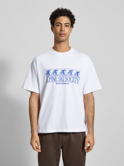 Männermodel trägt T-Shirt mit Label-Print von ANOTHER COTTON LAB, Reine Baumwolle, weiß mit blauem Print, Lockerer-Schnitt, Gerippter Rundhalsausschnitt, Rückenlänge bei Größe M 73 cm, Ganzkörperansicht von vorne.