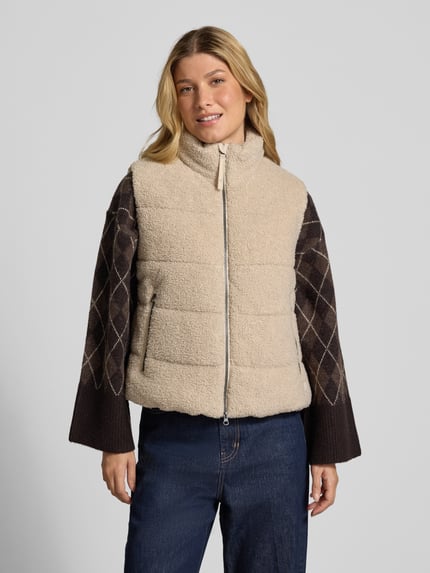 Modelka kobieta nosi Steppweste aus Teddyfell mit Zweiwege-Reißverschluss od camel active, beż, krój regular fit, stójka, kieszenie na zamek, długość pleców w rozmiarze 36: 55 cm, szerokość ramion w rozmiarze 36: 40 cm, szerokość w rozmiarze 36: 52 cm, widok całej sylwetki z przodu.