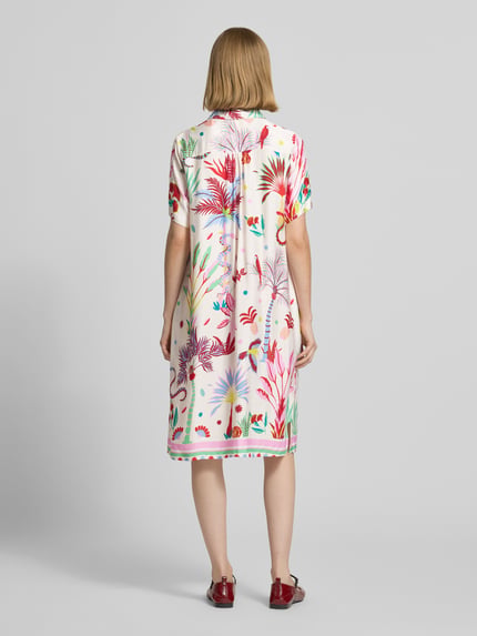 Vrouwenmodel draagt 'Knielanges Hemdblusenkleid aus Viskose mit Knopfleiste' van Milano Italy, wit met kleurrijke bloemenprint, loose fit, knielengte, overhemdkraag, korte knoopsluiting, ruglengte maat 36: 97 cm, borstwijdte maat 36: 52 cm, mouwlengte maat 36: 25 cm. Ganzlichaamsaanzicht van achteren.