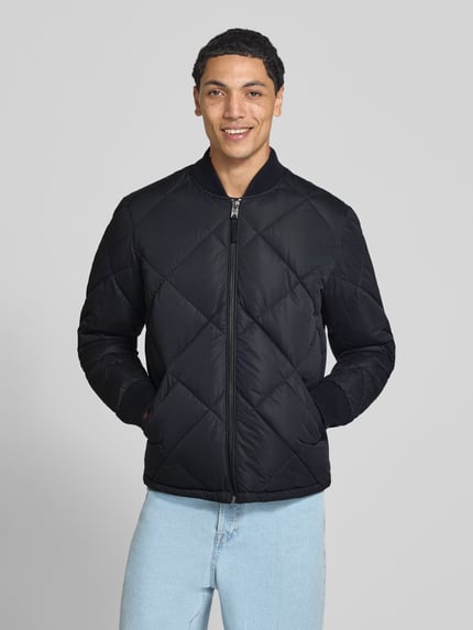 Männermodel trägt Regular Fit Steppjacke mit Stehkragen van Superdry in zwarte Stiksels look met steekzakken, elastische mouwboorden en ritssluiting, Ruglengte maat M 69 cm, schouderbreedte 46 cm, borstwijdte 58 cm, mouwlengte 59 cm, Ganzkörperansicht van voren.