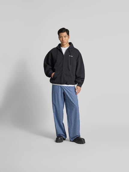 Männermodel trägt Pyjama-Hose aus reiner Baumwolle von VETEMENTS, Relaxed-Fit, kariertes Blau-Weiß-Muster, elastischer Bund, seitliche Eingrifftaschen, Label-Detail, Ganzkörperansicht von vorne.