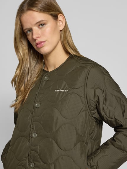 Kobiece model prezentuje kurtkę pikowaną o kroju regular fit z okrągłym dekoltem, wyhaftowane logo, listwa guzikowa, Carhartt Work In Progress Regular Fit Steppjacke mit Label-Stitching Model 'Skye Liner', długość tyłu w rozmiarze S: 61 cm, szerokość ramion w rozmiarze S: 41 cm, szerokość w rozmiarze S: 54 cm, długość rękawów w rozmiarze S: 61 cm, zbliżenie na górną część ciała.