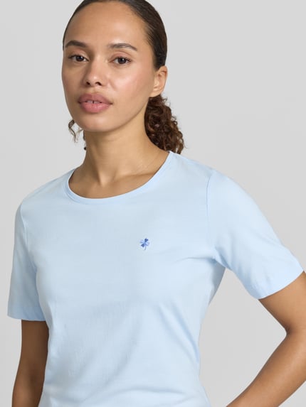 Modelka kobieca nosi T-Shirt mit Motiv-Stitching und Rundhalsausschnitt od Christian Berg Woman, czysta bawełna, krój regular fit, jasnoniebieski kolor, delikatna tekstura, haftowany motyw z przodu, widok na górną część tułowia z przodu.