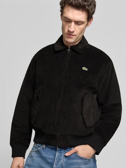 Männermodel trägt Bomberjacke mit Cordstruktur von LACOSTE in Schwarz, sichtbares Logo-Patch, seitliche Eingrifftaschen, Ganzkörperansicht von vorne.