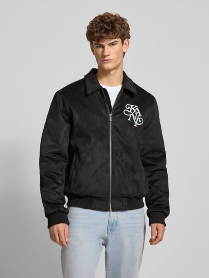 Männermodel trägt Jacke in Leder-Optik mit Label-Stitching von KARL KANI, Regular-Fit, Umlegekragen, Reißverschluss, schwarz, Ganzkörperansicht von vorne. Rückenlänge 68 cm, Schulterbreite 49 cm, Brustweite 59 cm, Ärmellänge 70 cm.