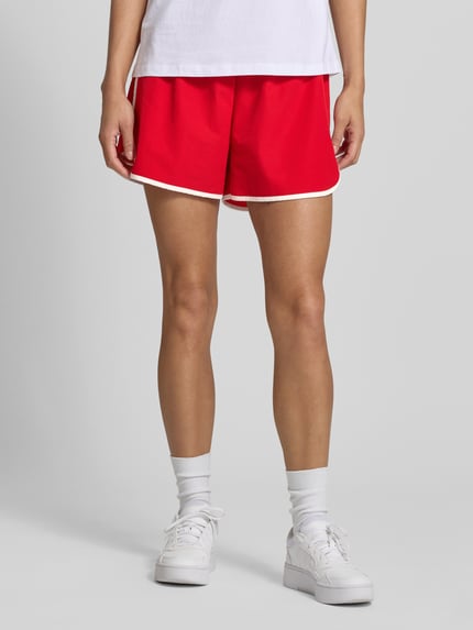 Frauenmodel trägt Track Shorts mit elastischem Bund und Label-Detail von Karo Kauer in Rot mit weißer Kante, Material-Mix mit Stretch-Anteil, Loose-Fit, Bundweite 36 cm, Innenbeinlänge 9 cm, Nahaufnahme Oberkörper.