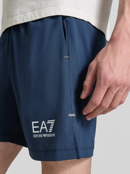 Męski model nosi Shorts mit Logo und elastischem Bund von EA7 Emporio Armani, granatowe, mieszanka materiałów z dodatkiem streczu, krój regular fit, logo, elastyczny pas, szerokość w rozmiarze M: 39 cm, długość wewnętrzna nogawki: 18 cm, zbliżenie na prawą nogawkę.