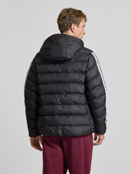 Männermodel trägt schwarze Regular Fit Steppjacke mit Kapuzenkragen von adidas Sportswear, weiße Streifen, Ganzkörperansicht Rückansicht.