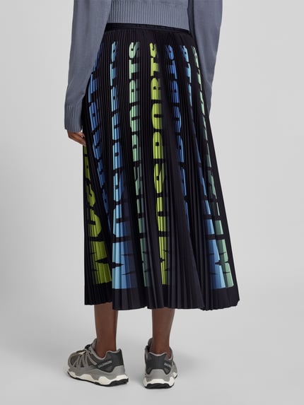 Frauenmodel trägt Midirock in Plissee-Optik von Marc Cain, schwarze Grundfarbe, bunte Schrift, elastischer Bund, Ganzkörperansicht Rückansicht.