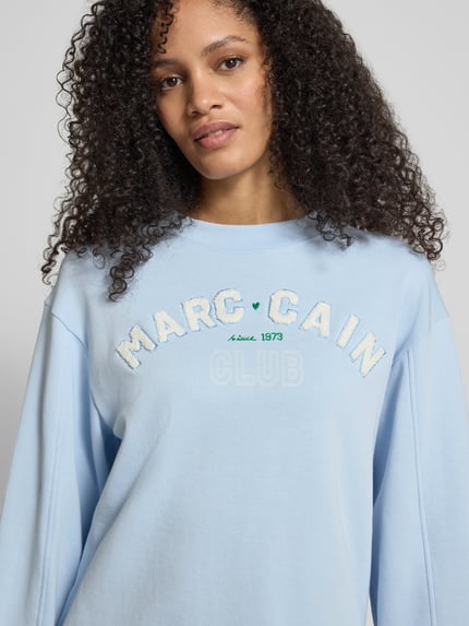 Frauenmodel trägt Relaxed Fit Sweatshirt mit Logo-Detail von Marc Cain, hellblauer Baumwoll-Mix, Rundhalsausschnitt, Nahaufnahme Oberkörper.