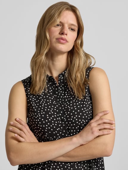 Vrouwenmodel draagt Ausgestelltes Hemdblusenkleid aus reiner Viskose Modell 'THEA' van Only met all-over motief, mouwloos, knoopsluiting, overhemdkraag, V-hals, uitlopend, minilengte, zwarte stof met witte stippen, gladde textuur, detailopname bovenlichaam.