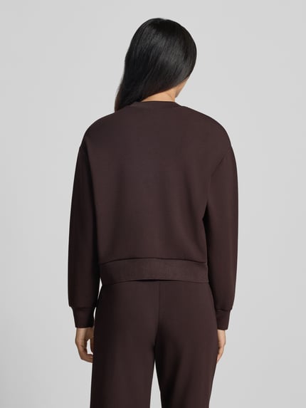 Frauenmodel trägt Sweatjacke mit Label-Print von Armani Exchange in Dunkelbraun, lockere Passform, glatte Textur, Ganzkörperansicht Rückansicht. Damen Sweatjacke von Armani Exchange, Viskose-Mix mit Stretch-Anteil, Zweiwege-Reißverschluss, seitliche Eingrifftaschen.