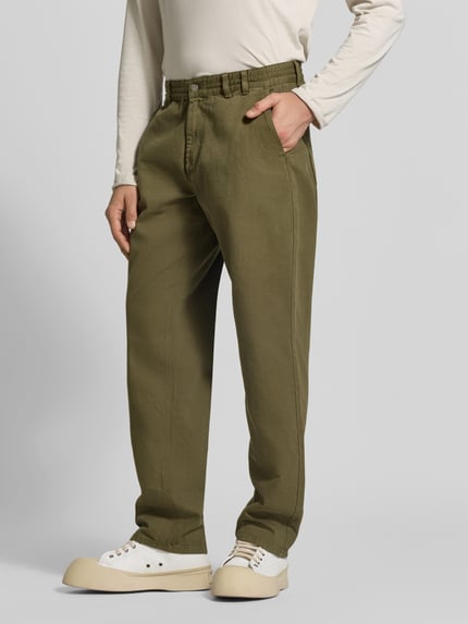 Männermodel trägt Stoffhose mit Label-Patch von Forét aus reiner Baumwolle, olivgrün, gerader Schnitt, Ganzkörperansicht von vorne.