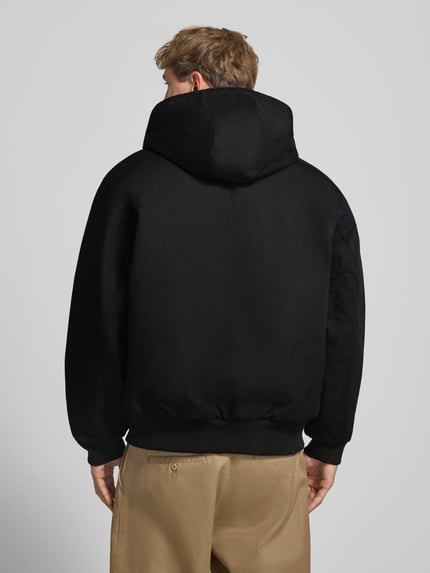 Männermodel trägt Jacke mit Label-Patch und Känguru-Tasche von Carhartt Work In Progress in Schwarz, glatte Baumwollstruktur, Ganzkörperansicht Rückansicht.