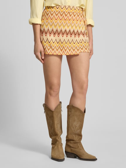 Kobiece model prezentuje spódnica mini 'Minirock im Stufen-Look' od Gina Tricot, mieszanka materiałów z dodatkiem streczu, linia a, efekt stopniowania, elastyczny pas, szerokość w rozmiarze S: 30 cm, całkowita długość: 37 cm, żółte, pomarańczowe i czerwone zygzaki, tekstura gładka, widok całej sylwetki z przodu.