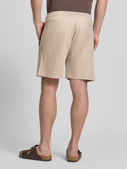 Mężczyzna model nosi Regular Fit Shorts mit Tunnelzug Modell 'JUANITO' von JOOP! Collection z bawełny, kolor beżowy, tekstura prążkowana, szerokość w rozmiarze M: 43 cm, długość wewnętrzna nogawki w rozmiarze M: 17 cm, widok z tyłu pełnej sylwetki.