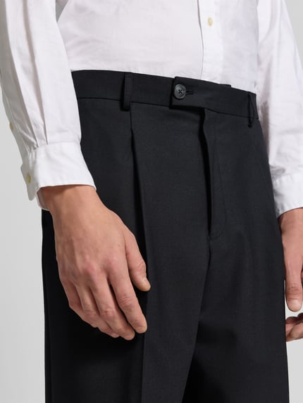 Männermodel trägt Straight Fit Stoffhose mit Bundfalten von ANNARR in Schwarz, glatte Textur, Nahaufnahme Oberkörper, Herrenmode-Kontext.