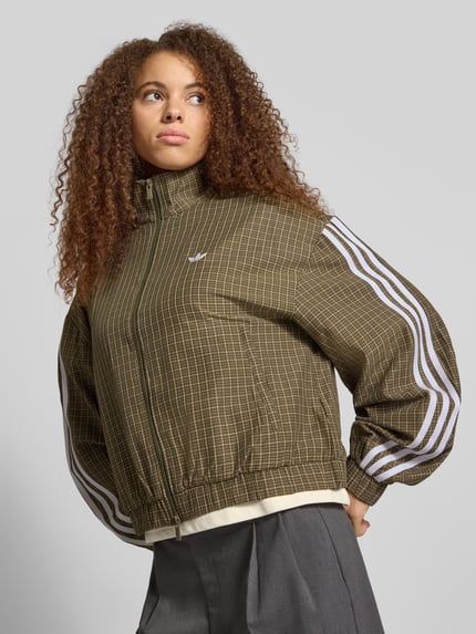 Modelka nosi Regular Fit Sweatjacke aus Viskose-Mix von adidas Originals, brązowy wzór kratka, białe paski, tekstura dzianinowa, całkowity widok z przodu.