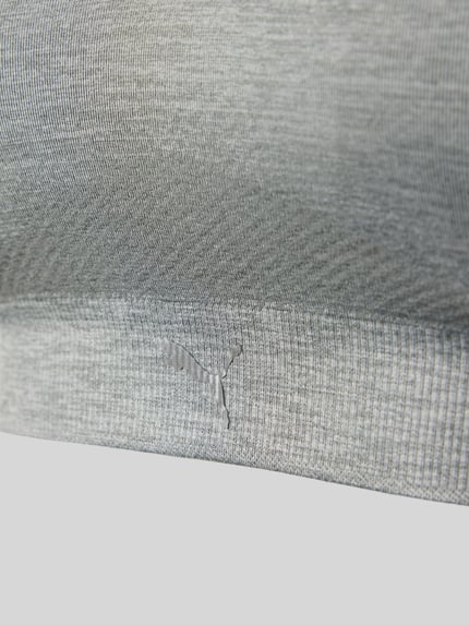 Frauenmodel trägt Triangel-BH mit Logo und gekreuzten Trägern von PUMA, grauer Material-Mix mit Stretch-Anteil, Logo, körpernahe Passform, Hakenverschluss, Nahaufnahme Oberkörper.