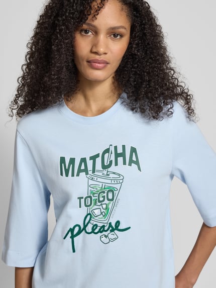 Frauenmodel trägt Slim Fit T-Shirt mit Motiv-Print von Marc Cain in Hellblau, reine Baumwolle, Halbarm, Rundhalsausschnitt, Nahaufnahme Oberkörper.