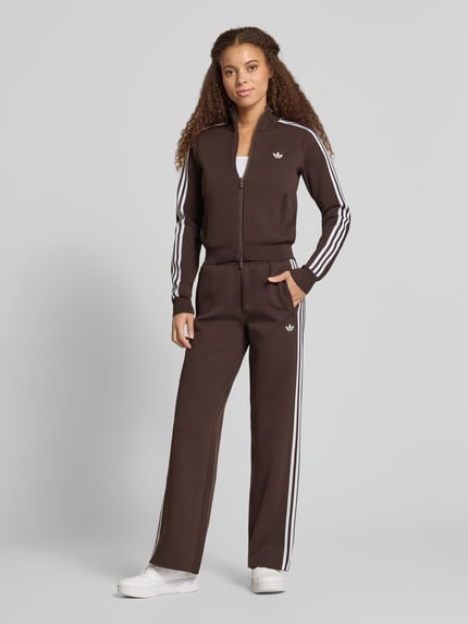 Frauenmodel trägt Regular Fit Sweathose mit Logo-Detail von Adidas Originals in Braun mit weißen Streifen, Teilungsnähten und Tunnelzug, Ganzkörperansicht von vorne.