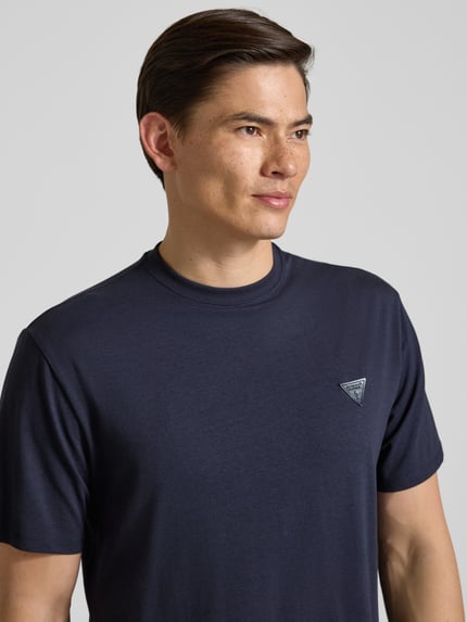 Männermodel trägt T-Shirt mit Logo und Rundhalsausschnitt von Guess, bawełna z domieszką modalu, krój relaxed fit, kolor granatowy, logo z przodu, widoczna tekstura materiału, Nahaufnahme Oberkörper.