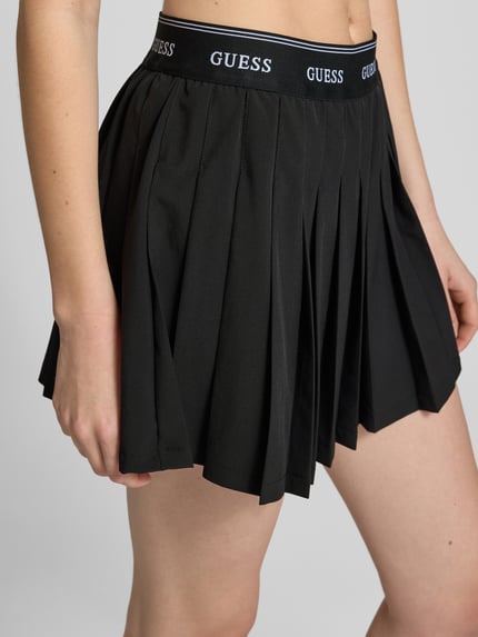 Frauenmodel trägt Faltenrock mit elastischem Bund Modell 'SELENE' von Guess Activewear, Minirock in Schwarz, Material-Mix mit Stretch-Anteil, A-Linie, Nahaufnahme Oberkörper. Bundweite 35 cm, Gesamtlänge 36 cm bei Größe S.