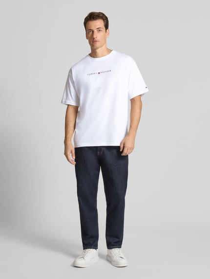 Męski model nosi Relaxed Fit T-Shirt aus reiner Baumwolle von Tommy Hilfiger, biały kolor, logo na piersi, krój relaxed fit, okrągły dekolt, krótki rękaw, czysta bawełna, długość tyłu w rozmiarze M: 73 cm, szerokość w rozmiarze M: 56 cm, jednolita faktura, widok całej sylwetki z przodu