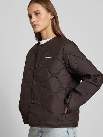 Frauenmodel trägt Steppjacke mit Logo-Stitching Modell 'Skye' von Carhartt Work In Progress in Dunkelbraun, sichtbar glatte Textur, Nahaufnahme Oberkörper. Rückenlänge 60 cm, Schulterbreite 42 cm, Brustweite 53 cm, Ärmellänge 62 cm.