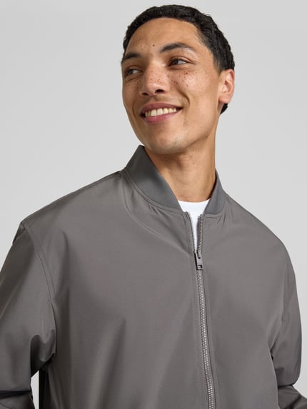 Męski model nosi bomberkę ze stójką model 'CHARGE' od Jack & Jones, krój regular fit, zamek błyskawiczny, szara, zbliżenie na górę ciała. Długość tyłu w rozmiarze M: 67 cm, szerokość ramion: 53 cm, szerokość klatki piersiowej: 62 cm, długość rękawów: 65 cm.